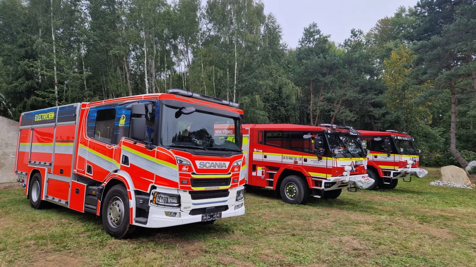 Drei Feuerwehrfahrzeuge, darunter ein elektrisches Scania-Modell, stehen auf einer Grünfläche vor einem Wald bei einer Hausmesse