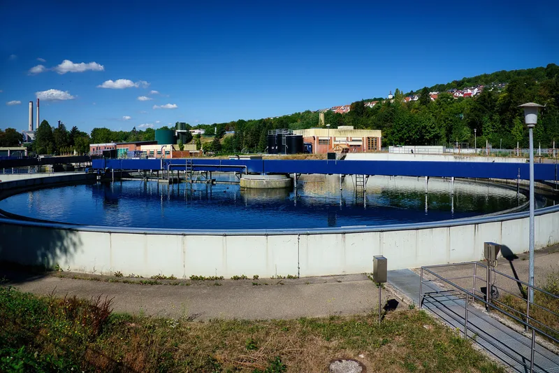 OT: Blaues Räumerbecken einer Kläranlage mit Betriebsgebäuden vor einer bebauten Hügellandschaft unter blauem Himmel