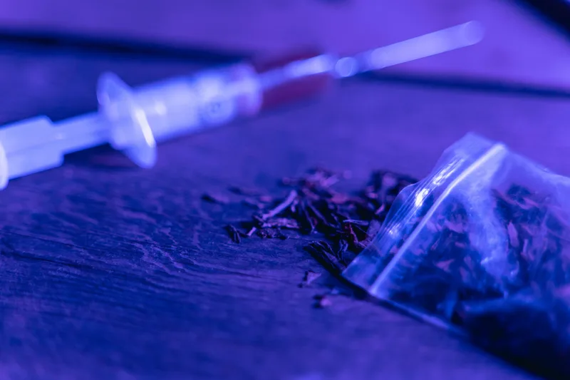 BKA: Einwegspritze mit Flüssigkeit und offener Beutel mit getrockneten Pflanzenteilen unter blauem Licht auf einer Holzoberfläche