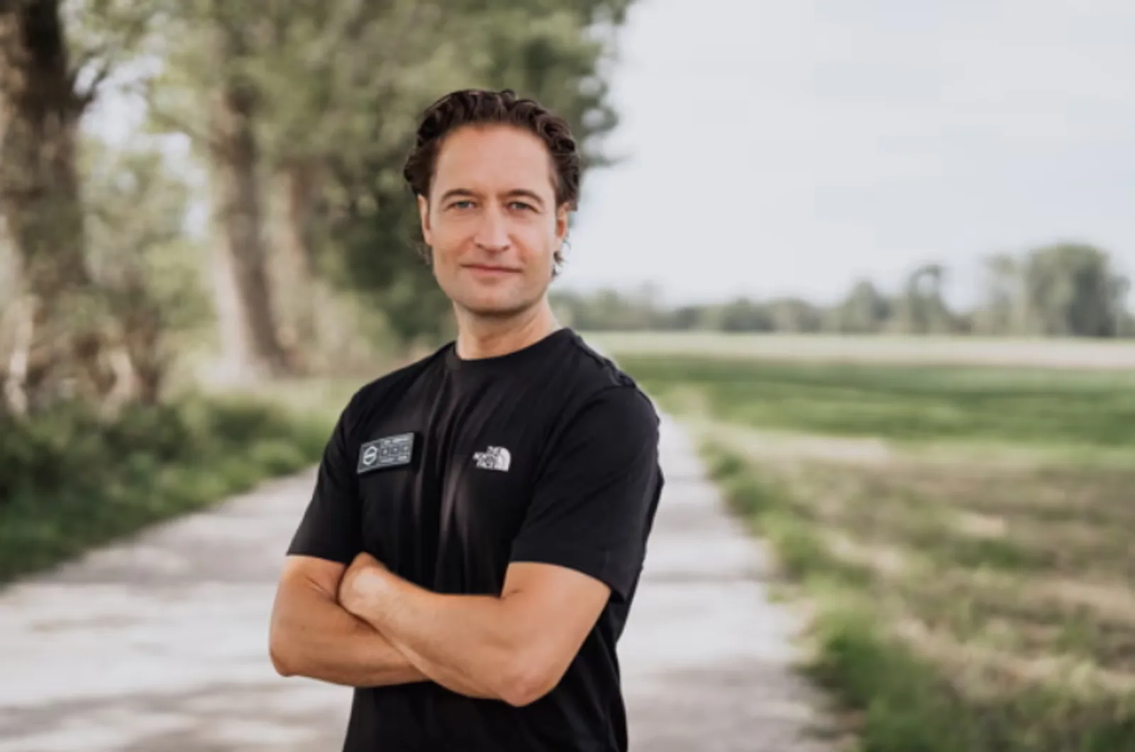 Florian Taitsch von ARX Robotics steht mit verschränkten Armen in einem schwarzen T-Shirt auf einem ländlichen Feldweg