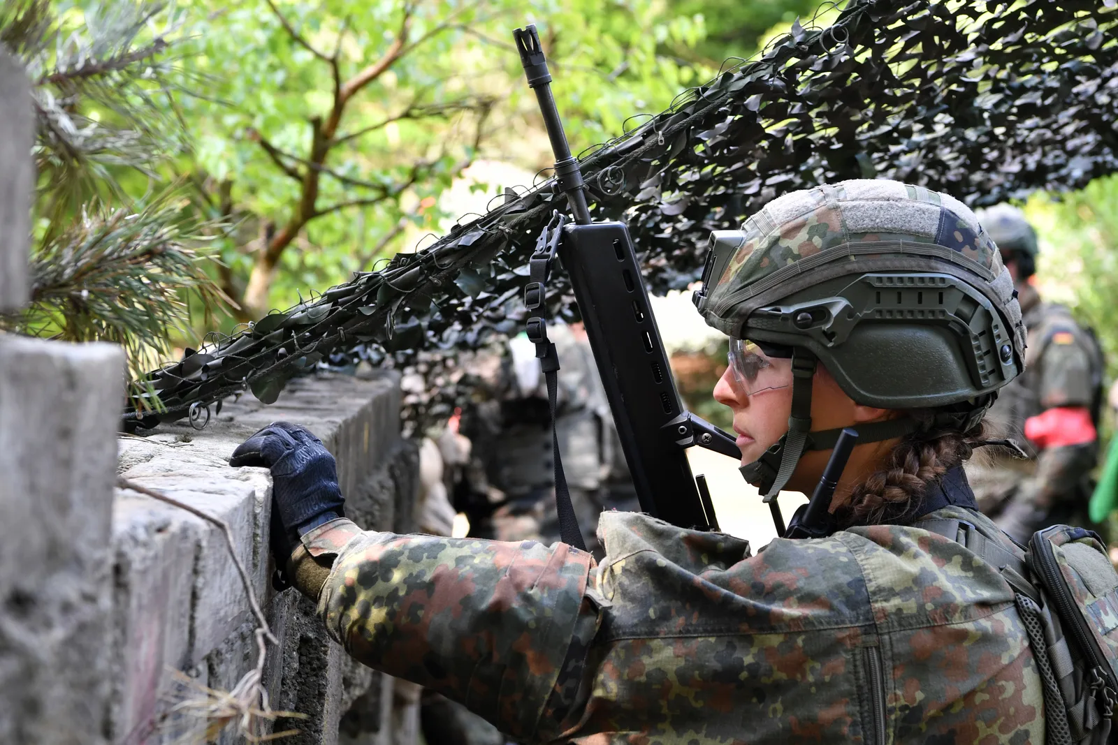 Bundeswehr-Soldatin in Flecktarn sichert mit einem G36 hinter einer Mauer unter einem Tarnnetz bei der Übung Vigilant Roland
