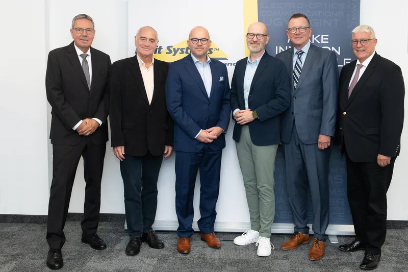 Sechs Männer in Anzügen posieren vor Werbebannern von Elbit Systems Deutschland bei einer Unternehmenspräsentation