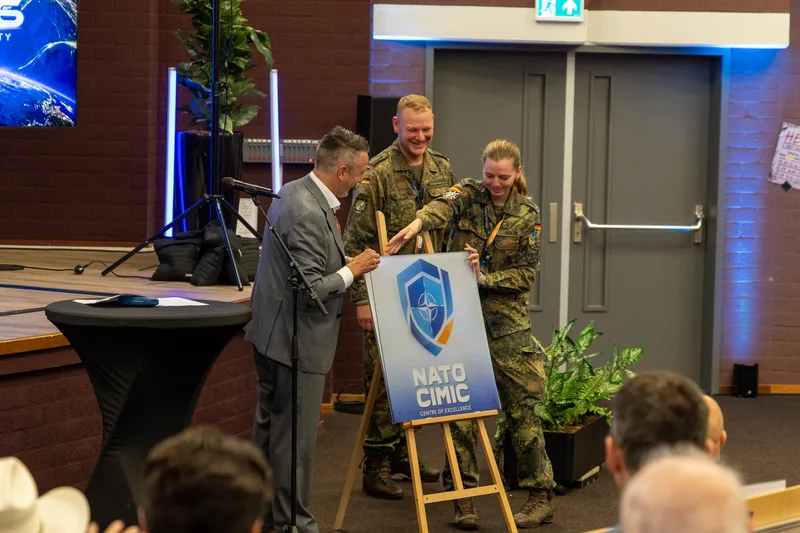 Ein Mann im Anzug sowie zwei Bundeswehrsoldaten präsentieren ein Schild mit dem Logo des NATO-CIMIC-Kompetenzzentrums auf einer Bühne