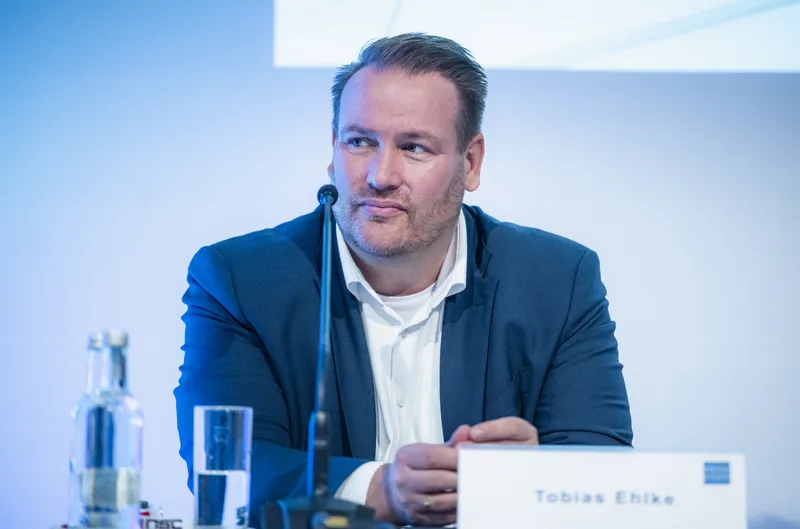 Tobias Ehlke vom CPM Verlag sitzt bei einer Vortragsveranstaltung an einem Tisch mit Mikrofon, Namensschild und Wasserglas