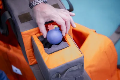 Hand platziert ein blaues, bombenförmiges Übungsobjekt in einer gepolsterten, grau-orangenen Transporttasche für Sicherheitsausrüstung