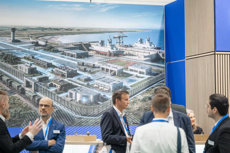 Besucher im Gespräch an einem Messestand vor einer Grafik mit militärischer Infrastruktur, Hafen und einem Airbus A400M