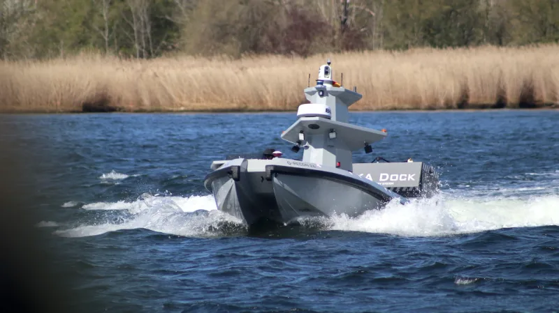 Unbemanntes Oberflächenboot Q-RECON 24 mit Sensormast auf einem Gewässer vor einer bewachsenen Uferlinie