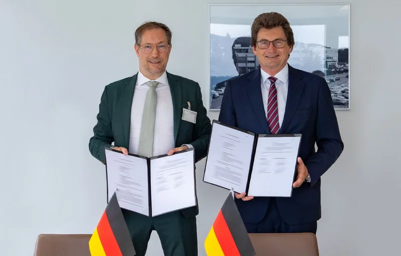 Zwei Vertreter präsentieren unterzeichnete Dokumente einer Absichtserklärung mit dem Unternehmen FLANQ vor zwei deutschen Tischflaggen