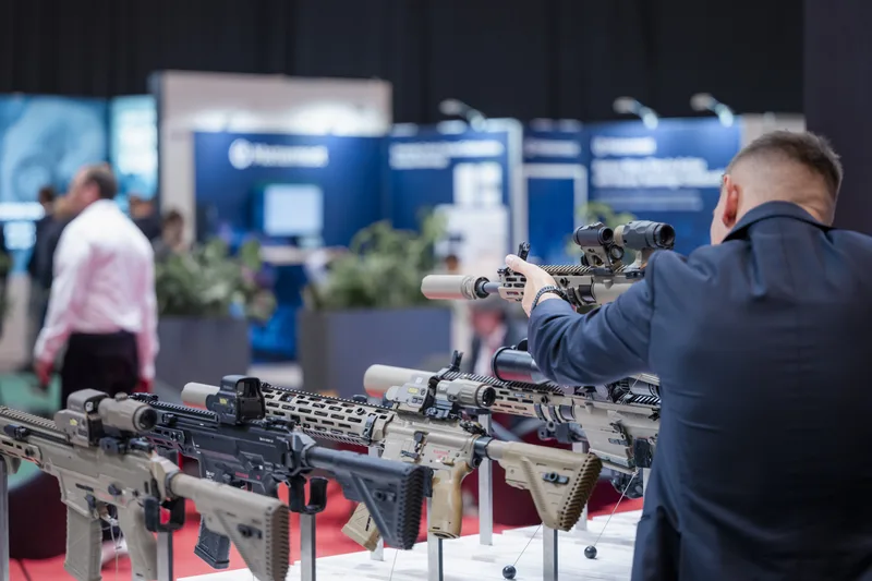 Besucher visiert auf einer Fachmesse ein Heckler & Koch Sturmgewehr an, im Vordergrund HK437-Modelle, im Hintergrund Patria-Stand