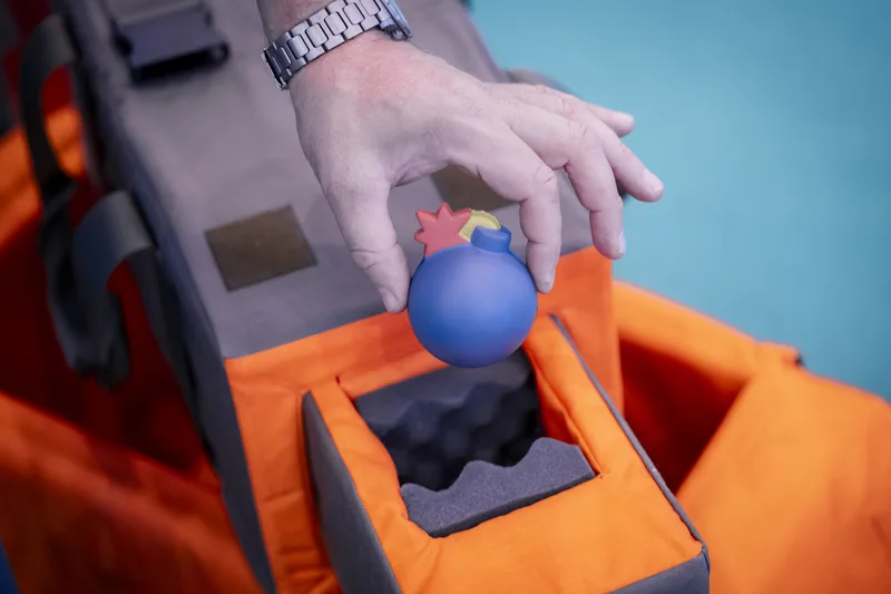 Hand legt einen blauen, bombenförmigen Übungsgegenstand in eine gepolsterte orange-graue Transporttasche