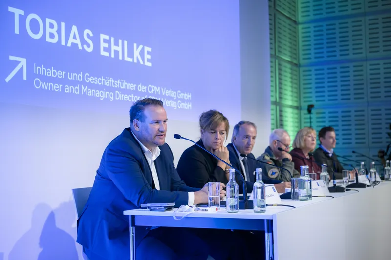 Tobias Ehlke von der CPM Verlag GmbH spricht bei einer Podiumsdiskussion vor Publikum, darunter ein Soldat der Bundeswehr