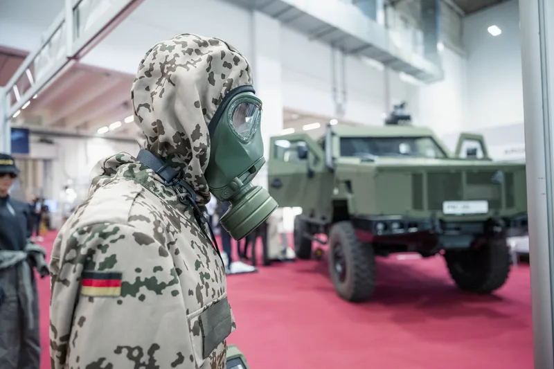 Bundeswehr-Uniform im Wüstentarn mit ABC-Schutzmaske vor einem geschützten Radfahrzeug vom Typ Enok auf einer Fachmesse