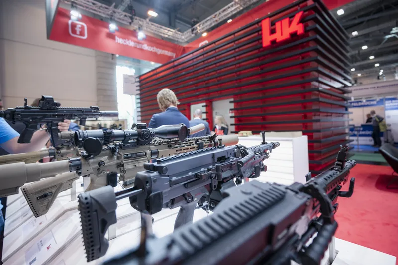 Sturmgewehre und Maschinengewehre von Heckler & Koch am Messestand auf einer Fachmesse mit Besuchern im Hintergrund