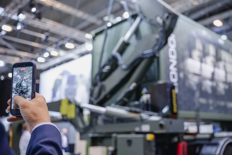 Besucher filmt mit Smartphone ein militärisches Wechselladesystem auf einem Lkw-Fahrgestell in einer Messehalle