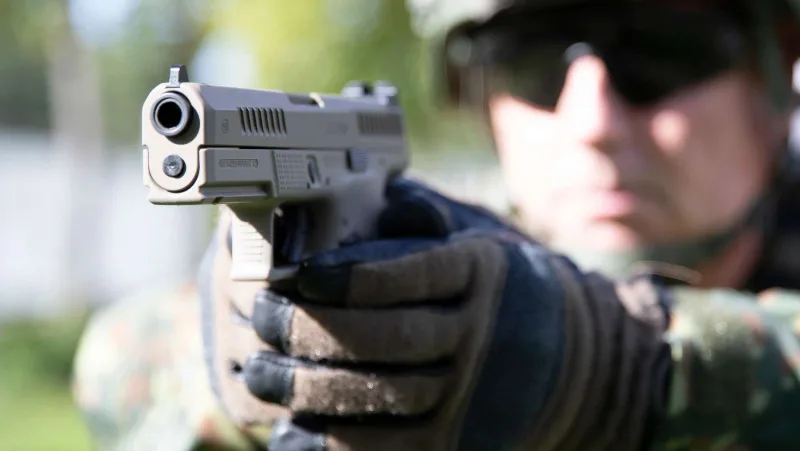 Soldat in Flecktarn-Uniform mit Gefechtshelm und Schutzbrille zielt mit einer sandfarbenen CZ P-10 Pistole in Richtung Kamera