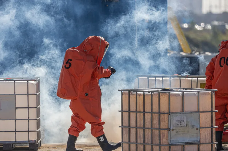 Einsatzkräfte in orangefarbenen Chemikalienschutzanzügen bei einer Gefahrgutübung zwischen Gittertanks in dichtem Rauch