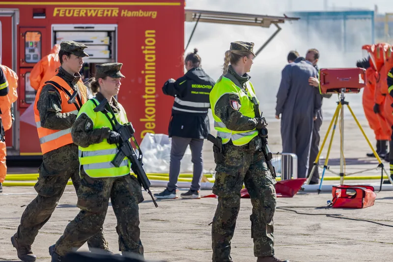 Bundeswehrsoldaten mit G36 sichern eine Dekontaminationsübung mit Einsatzkräften der Feuerwehr Hamburg und Chemikalienschutzanzügen