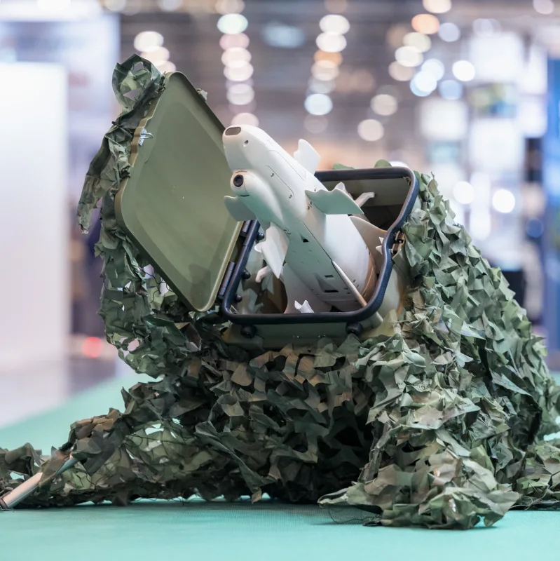 Weiße Drohne mit klappbaren Flügeln und optischem Sensor in einem mit Tarnnetz bedeckten Transportbehälter auf einer Fachmesse