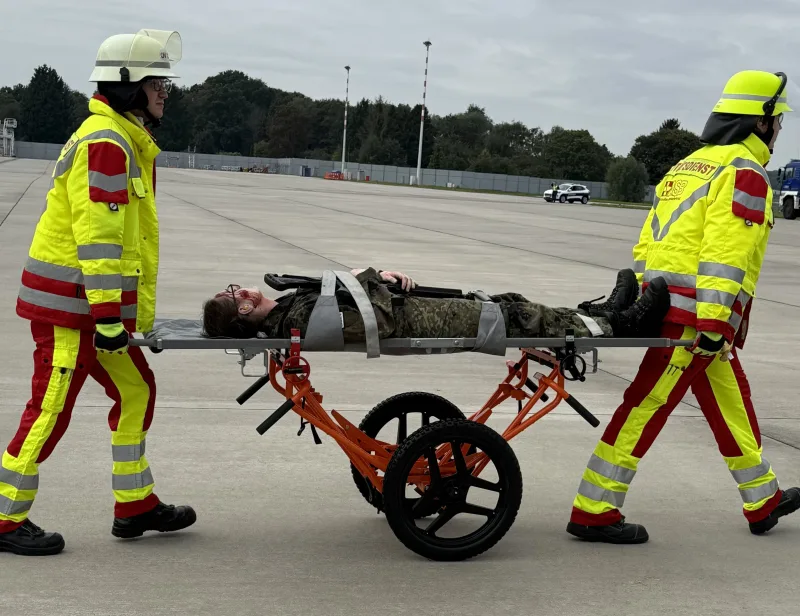 Red Storm Charlie: Zwei Einsatzkräfte des Arbeiter-Samariter-Bundes transportieren einen verletzten Soldaten auf einer Krankentrage über ein Flugfeld