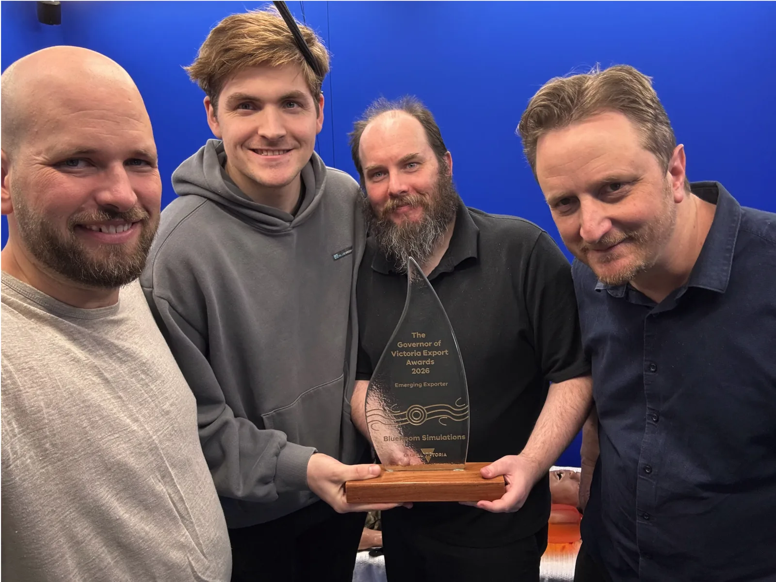 Vier Mitarbeiter von BlueRoom Simulations präsentieren eine Auszeichnung der Governor of Victoria Export Awards vor blauem Hintergrund