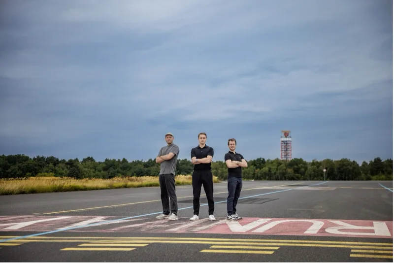 Drei Männer stehen mit verschränkten Armen auf einer Flugplatz-Landebahn vor einem Kontrollturm und Waldgebiet