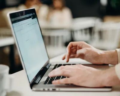 NoSpamProxy: Hände tippen auf einer Laptop-Tastatur an einem hellen Schreibtisch vor unscharf erkennbaren Personen im Büro