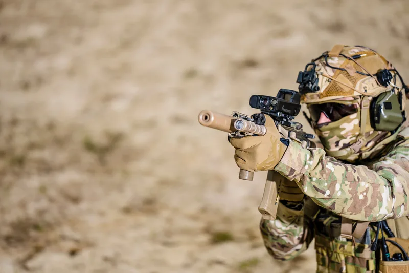 Soldat in Tarnkleidung zielt mit einem Sturmgewehr inklusive Smart Shooter SMASH Feuerleitsystem und Schalldämpfer im sandigen Gelände