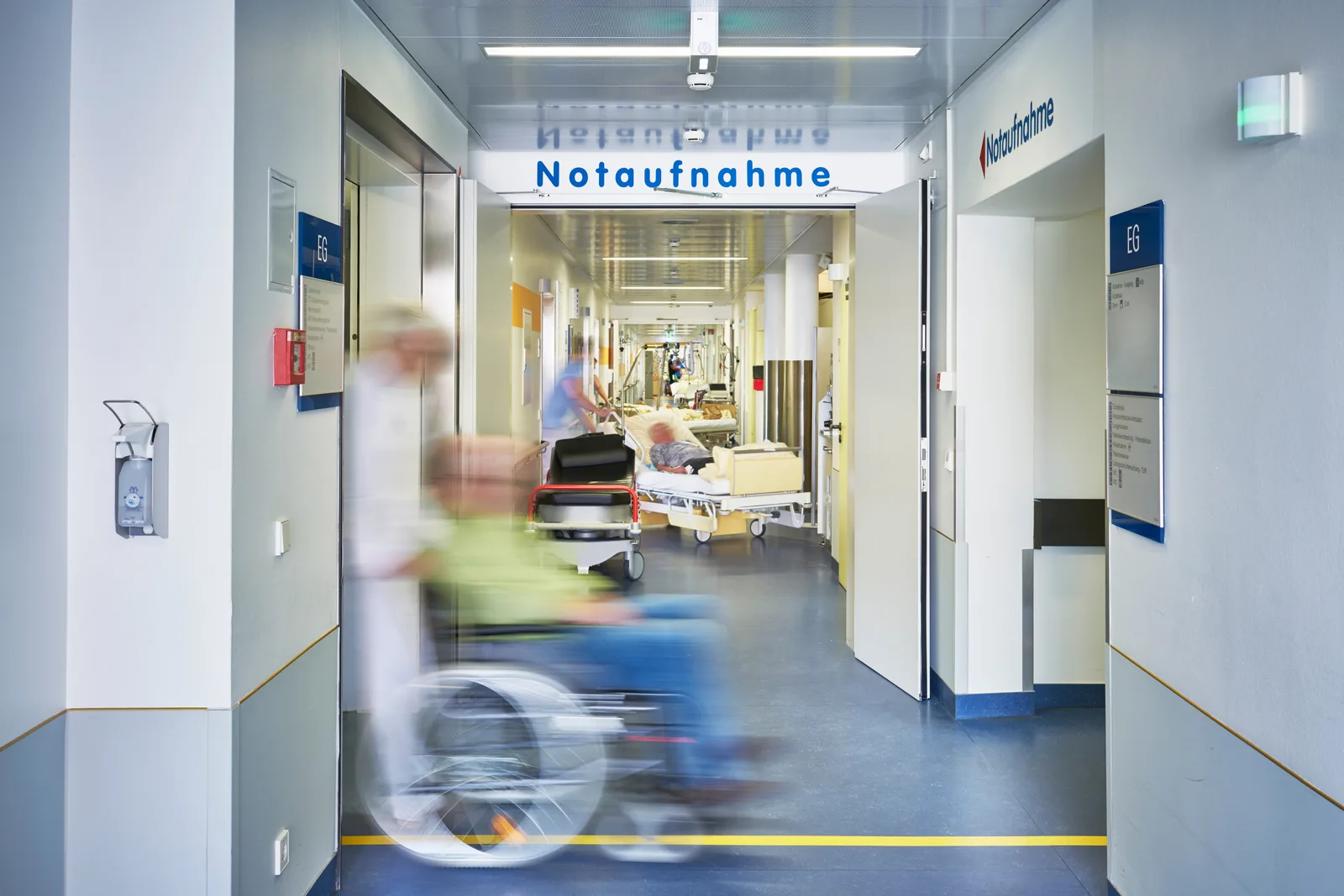 Patient im Rollstuhl auf dem Flur einer Notaufnahme mit medizinischem Personal und Patienten in Krankenbetten im Hintergrund