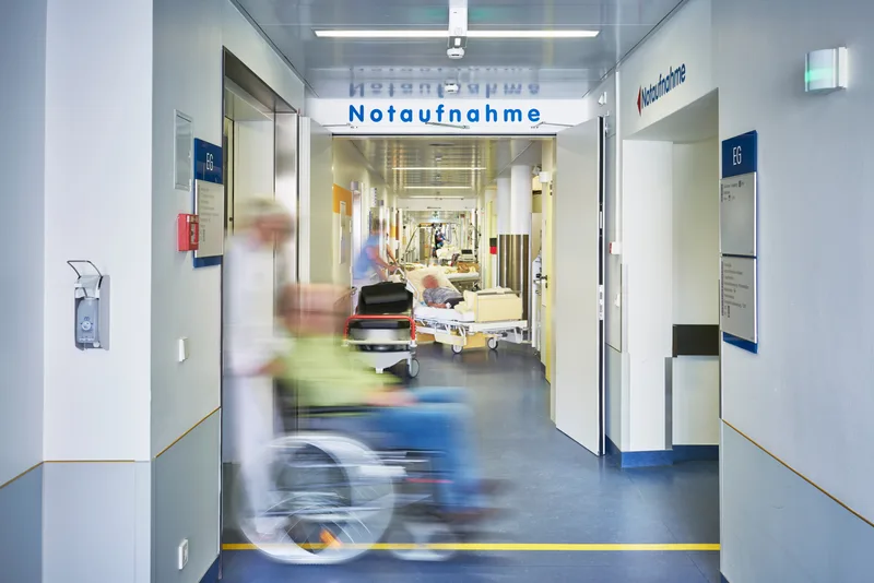 Patient im Rollstuhl auf dem Flur einer Notaufnahme mit medizinischem Personal und Patienten in Krankenbetten im Hintergrund