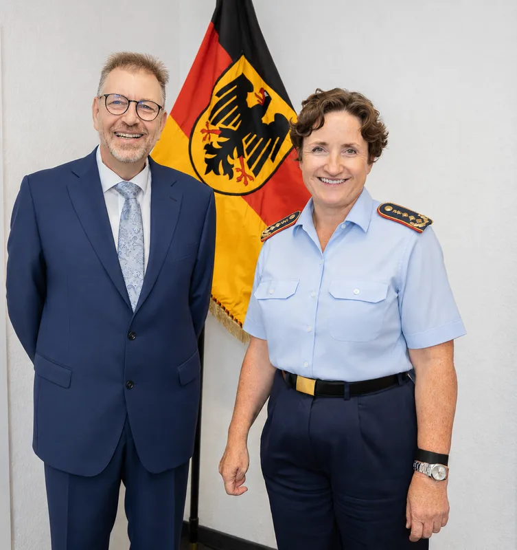 Generalstabsärztin der Bundeswehr in Uniform und ein Mann im blauen Anzug stehen lächelnd vor einer Bundesdienstflagge