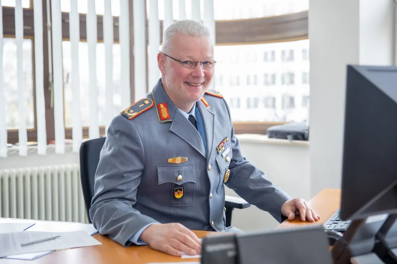 Generalarzt Dr. Ahrens in Bundeswehr-Dienstanzug an seinem Schreibtisch mit Dokumenten, Monitor und Tastatur im Büro
