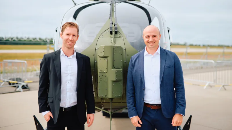Zwei Männer in Anzügen stehen vor der Front eines militärischen Airbus H145M Hubschraubers auf einem Flugplatz
