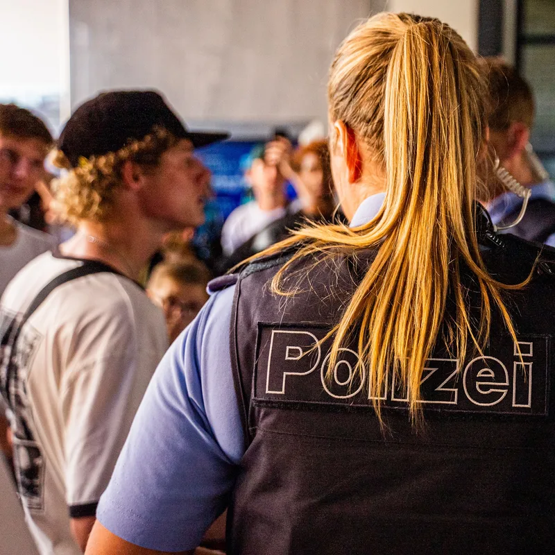 Polizeibeamtin in Schutzweste steht mit dem Rücken zur Kamera vor einer Gruppe junger Menschen bei einem Polizeieinsatz. Frauen bei der hessischen Polizei werden gewürdigt