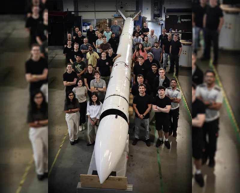 Mitarbeiter von HyImpulse posieren in einer Montagehalle neben der auf Gestellen gelagerten suborbitalen Rakete SR75