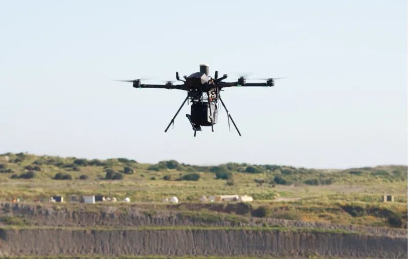 Unbemannte Multirotor-Drohne mit Sensornutzlast im Schwebeflug über einem weitläufigen, terrassierten Gelände