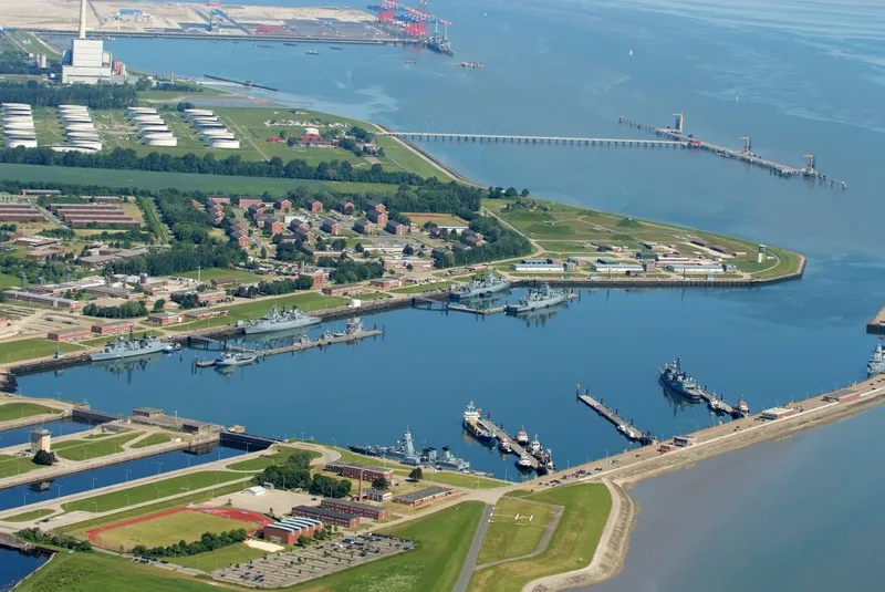 Luftaufnahme des Marinestützpunkts Heppenser Groden in Wilhelmshaven mit Fregatten an den Piers sowie Industrieanlagen im Hintergrund