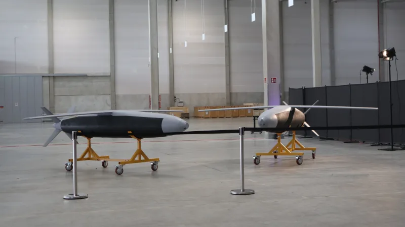 Zwei militärische Flugkörper auf gelben Transportgestellen in einer Industriehalle mit Studiobeleuchtung und Absperrband