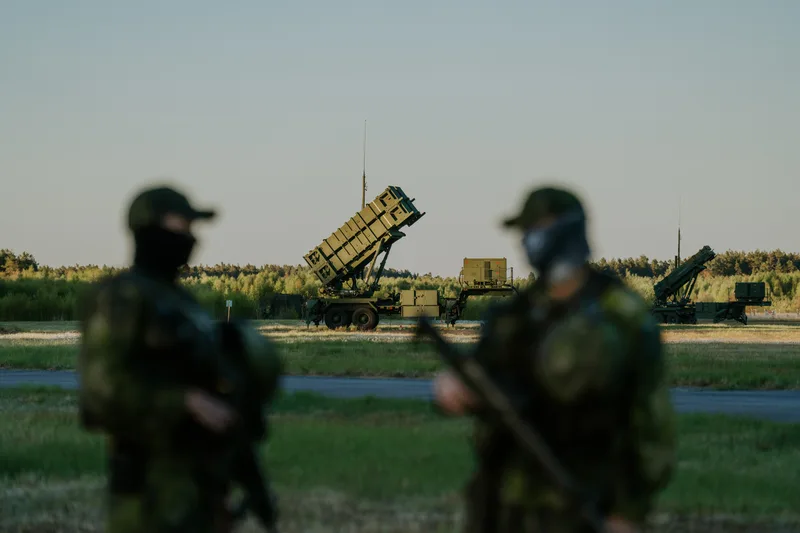 Zwei bewaffnete Soldaten in Flecktarn sichern vor einem in Feuerstellung befindlichen MIM-104 Patriot Flugabwehrsystem