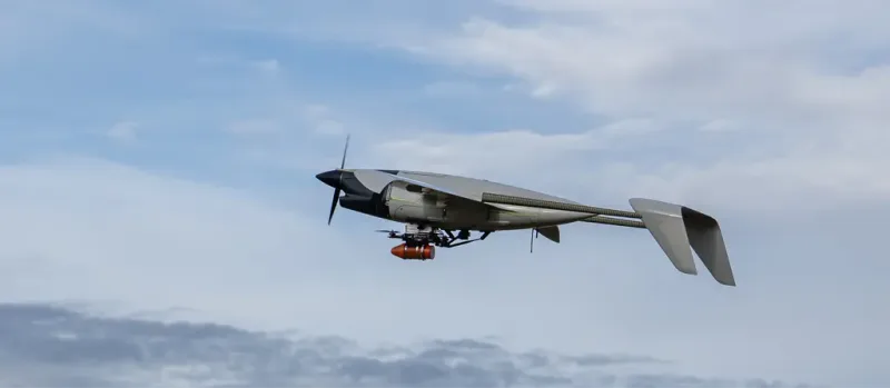 Starrflügeldrohne mit Frontpropeller und montiertem Multikopter als Außenlast im Flug vor bewölktem Himmel