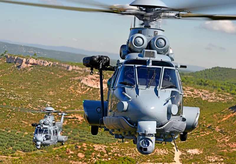 Zwei Airbus H225M Hubschrauber fliegen in Formation über hügeligem Gelände, die vordere Maschine mit Sensorsystem unter dem Bug