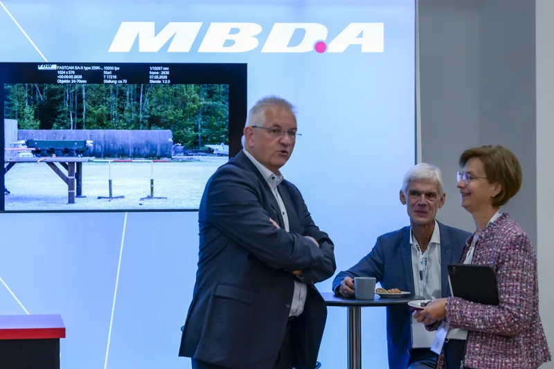Fachbesucher im Gespräch an einem Messestand von MBDA vor einem Bildschirm mit technischen Testaufnahmen