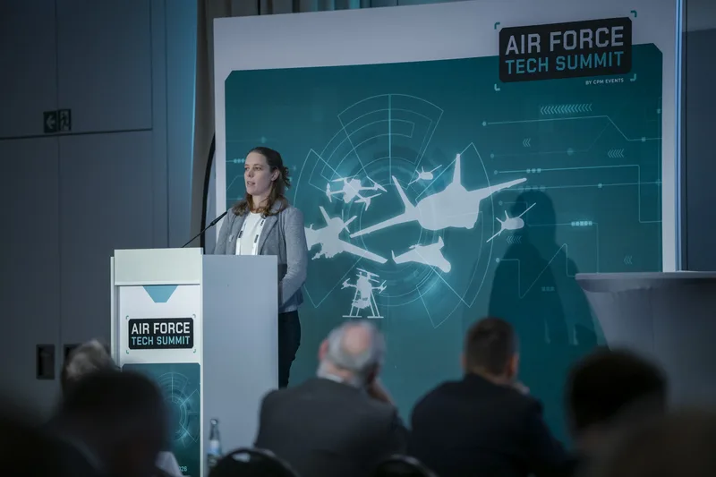 Referentin spricht am Rednerpult auf dem Air Force Tech Summit vor einer Bühnenrückwand mit Grafiken von Militärflugzeugen