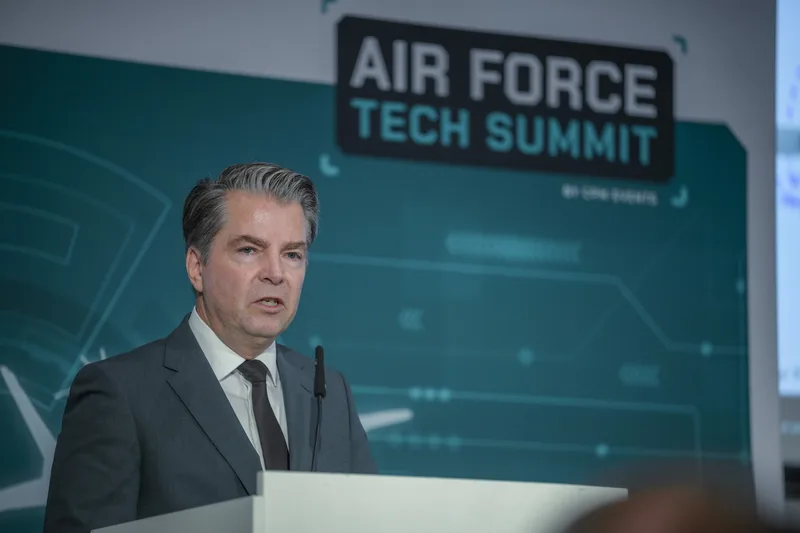 Referent spricht an einem Rednerpult auf dem Air Force Tech Summit vor einer gebrandeten Bühnenwand
