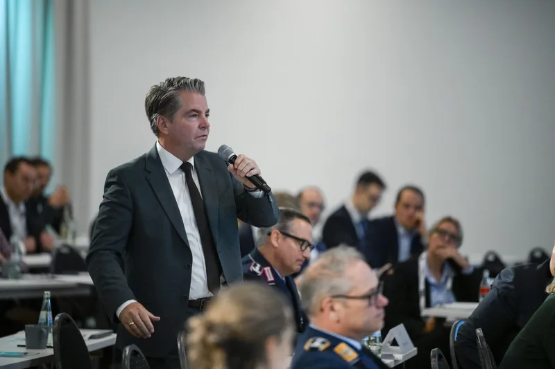 Ein Konferenzteilnehmer spricht mit einem Handmikrofon vor einem Publikum aus Zivilisten und Bundeswehrsoldaten in Dienstuniform