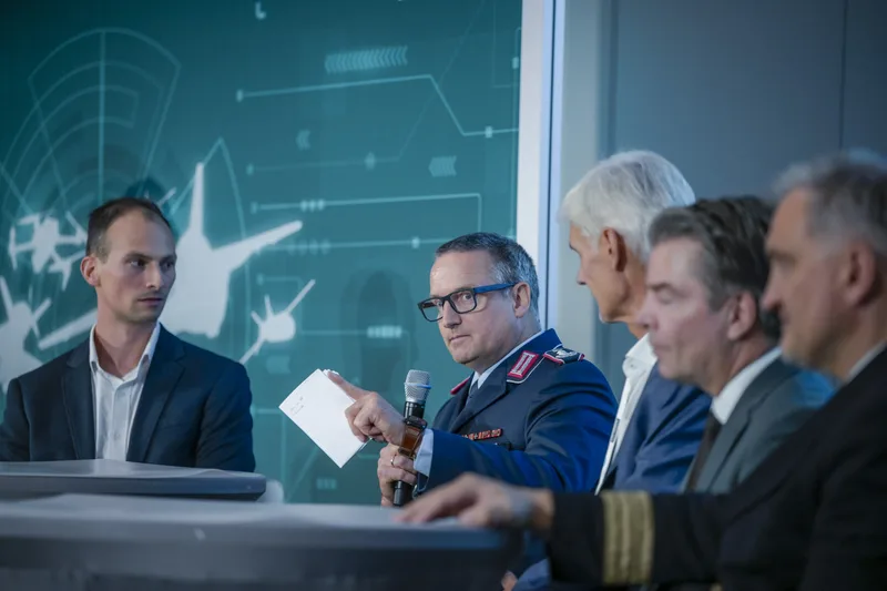 Luftwaffenoffizier der Bundeswehr spricht bei einer Podiumsdiskussion in ein Mikrofon vor einer Grafik mit Luftfahrzeugen und Radarsystemen