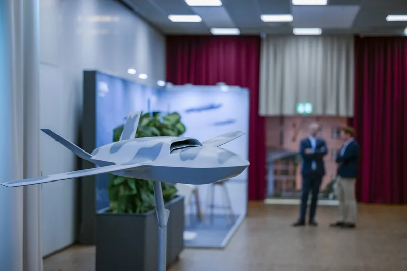 Modell einer getarnten Drohne auf einem Präsentationsständer vor Konferenzteilnehmern auf einer Wehrtechnikmesse