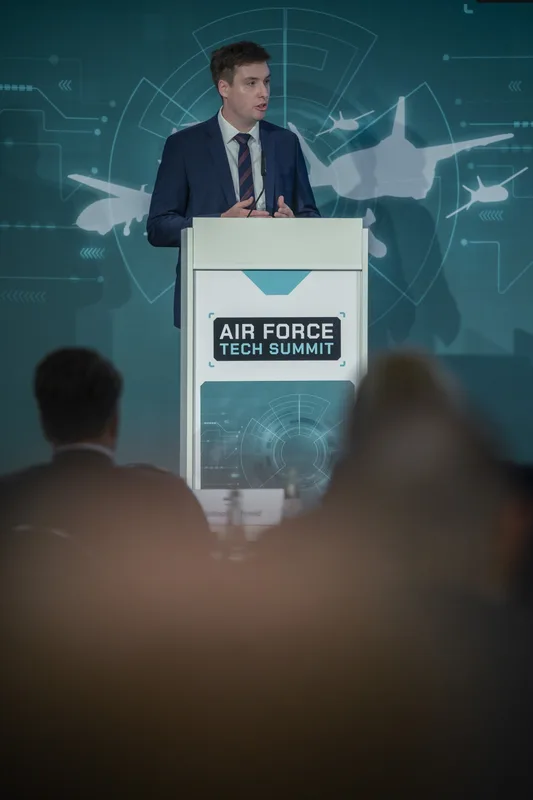 Referent spricht am Rednerpult auf dem Air Force Tech Summit vor einer Wand mit Grafiken militärischer Flugzeuge und Drohnen