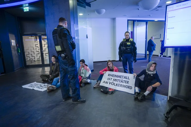Demonstranten bei einer Sitzblockade gegen Rheinmetall im Foyer eines Gebäudes, beobachtet von zwei Polizeibeamten