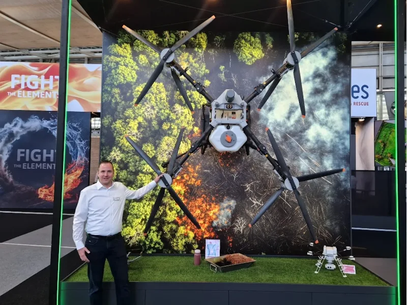 Aussteller präsentiert eine große DJI-Agras-Drohne an einer Waldbrand-Wand sowie ein kleineres Fluggerät auf einer Fachmesse