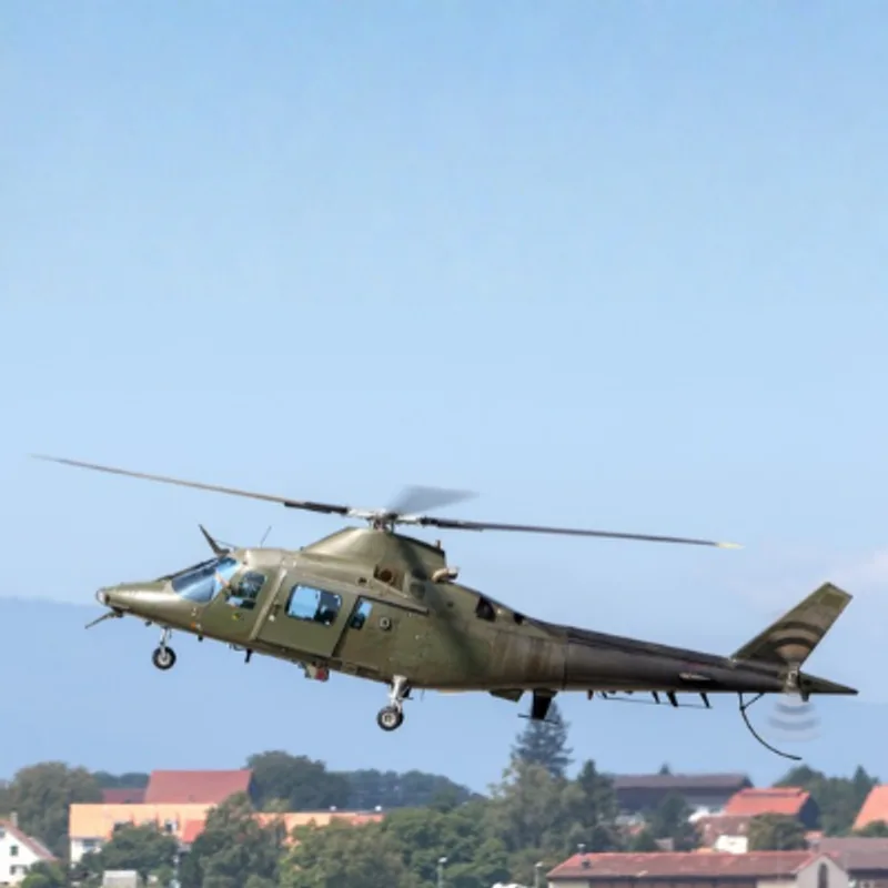 Militärischer Mehrzweckhubschrauber Leonardo AW109 mit ausgefahrenem Fahrwerk im Flug über einer bewaldeten Siedlung